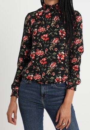 Femme portant un chemisier noir à fleurs à manches longues avec poignets smockés et col haut, associé à un jean en denim bleu.
