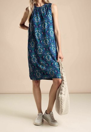 Kobieta ubrana w niebieską sukienkę bez rękawów o wzorze, sięgającą do kolan, białe tenisówki, trzyma dużą beżową dzianinową torbę typu tote.