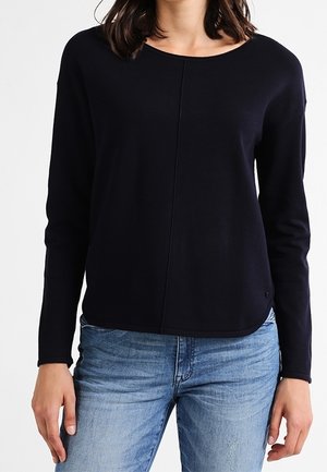 Femme portant un t-shirt noir uni à manches longues avec une couture centrale et un jean en denim bleu clair, visible des épaules aux hanches.