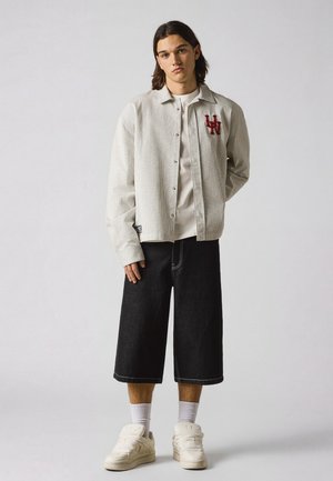 Ung mand med skulderlangt hår iført en ternet skjorte med knapper, hvid t-shirt, sorte shorts med vide ben, hvide sokker og hvide sneakers.