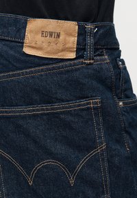 Dark blue denim jeans with orange stitching, back pocket, and a tan leather patch labeled "EDWIN" with Japanese text below.