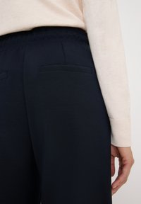 Marineblauwe broek met een gladde textuur, elastische tailleband en twee achterzakken. Het zicht bevat een mouw van een lichtgekleurde trui.