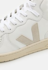 Vita lädersneakers med en beige mocka-logotyp, texturerad gummisula och vita snörningar. Har sömmar längs sidorna.