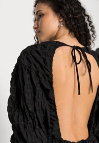 Vestido preto com um tecido texturizado e enrugado. Apresenta um decote profundo nas costas atado com uma fita e mangas bufantes com punhos elásticos.