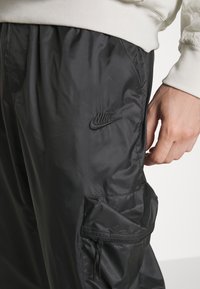 Černé nylonové cargo kalhoty s elastickým pasem, disponující boční kapsou a vyšitým logem Nike na stehně. Hladký povrch.