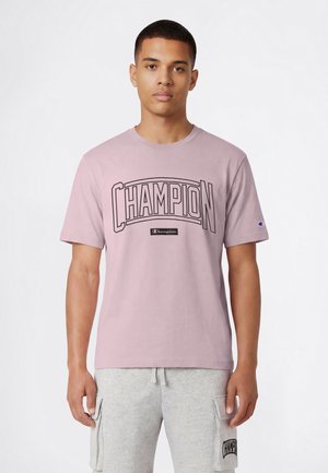 Młody mężczyzna w jasnofioletowej koszulce Champion i jasnoszarych szortach typu cargo stoi na tle białego tła.