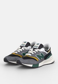 Šedé tenisky New Balance 997R s zelenými akcenty, vyrobené z materiálů mesh a semiš, bílé mezipodešve a černé podrážky. Obsahují žluté detaily.