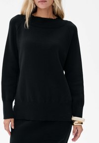 Zwarte gebreide sweater met een ruime halslijn, geribbelde kraag en lange mouwen. Heeft een rechte zoom en wordt gecombineerd met gestapelde armbanden.