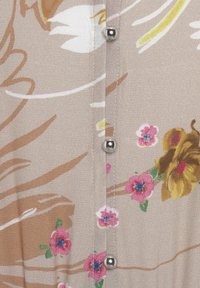 Blusa floral con una tela beige adornada con flores rosas, acentos marrones y botones plateados a lo largo del centro, creando un patrón texturizado.