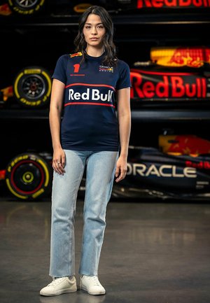 Granatowa koszulka Red Bull Racing z czerwonym i białym logo, w zestawieniu z jasnoniebieskimi dżinsami i białymi trampkami. W tle widać opony wyścigowe.