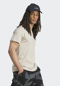 Camiseta de algodón beige de manga corta con un pequeño logo blanco, combinada con pantalones cortos de camuflaje gris oscuro y una gorra negra con un logo blanco.