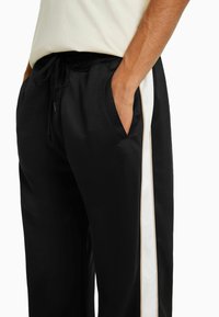 Black athletic pants with a white vertical stripe, Featuring a drawstring waist, side pockets, and smooth texture.