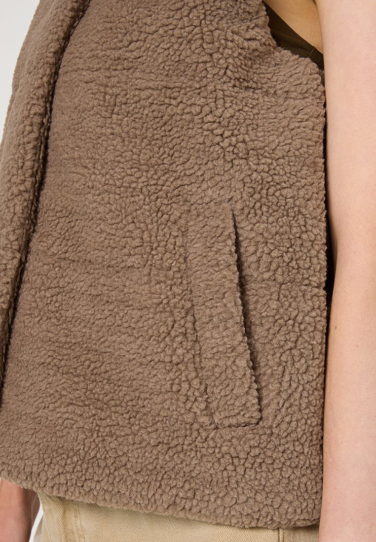 Gilet en polaire marron avec une surface douce et texturée. Dispose d'une poche plaquée à l'avant et d'un design sans manches.