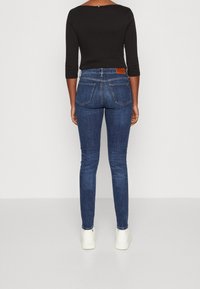 Vaqueros ajustados en denim azul oscuro con cintura media, diseño de cinco bolsillos y una etiqueta de cuero marrón en la parte posterior. Combinados con zapatillas blancas.
