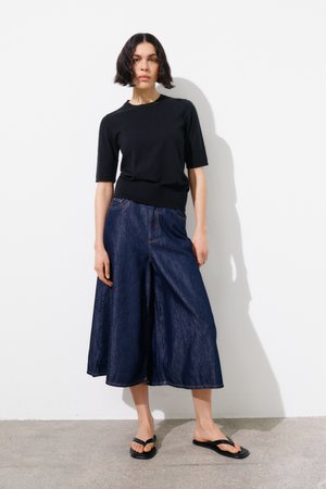 Nő áll fehér fal előtt, fekete rövid ujjú felsőt, széles sötét farmer culottes nadrágot és fekete papucsot visel.