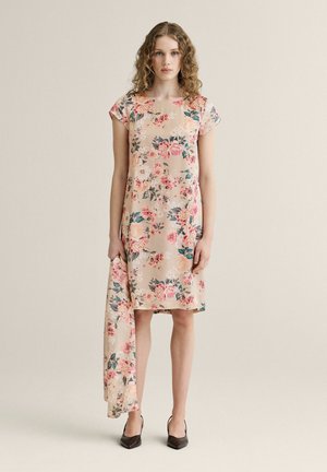 Kvinde iført en beige blomsterknælængde kjole, der holder matchende stof, iført mørke spidse sko, stående mod en ensfarvet neutral baggrund.