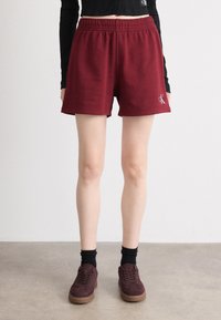 Burgundinės sportinės šortai su elastinga juosta ir CK logotipu. Derinami su burgundiniais sportiniais bateliais ir juodomis ilgojiomis kojinėmis, prieš neutralią fono spalvą.