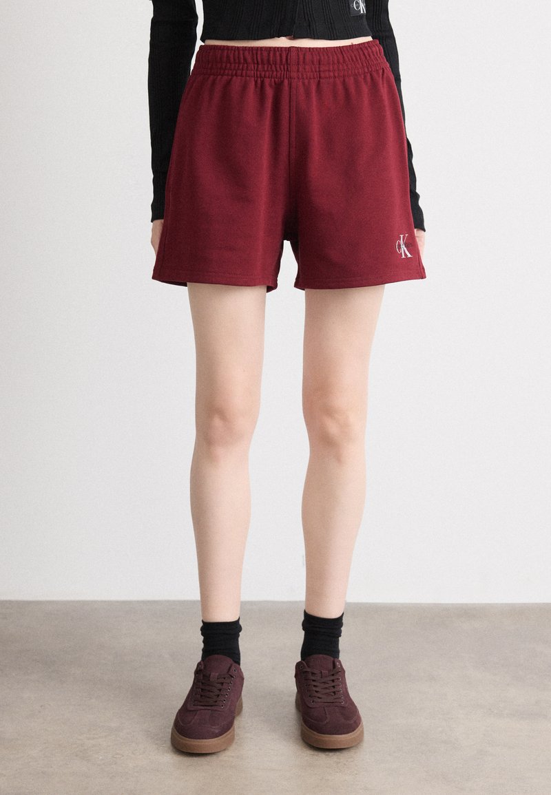 Shorts sportivi color marrone scuro con cintura elastica e logo CK. Abbinati a sneakers bordeaux e calzini neri, su uno sfondo neutro.