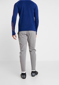 Top atlético de manga larga azul con acentos naranjas en las mangas, combinado con pantalones gris claro ajustados y zapatillas deportivas negras con suelas texturizadas.