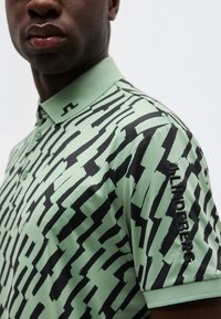 Άνδρας που φοράει ανοιχτό πράσινο πόλο με μαύρο αφηρημένο ζιγκ-ζαγκ μοτίβο και την επιγραφή "JL INDEPENDENCE" κεντημένη στο αριστερό μανίκι.