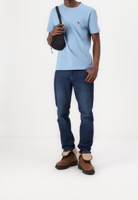 Hombre que lleva una camiseta azul clara, jeans azules, botas de borlas marrones, sosteniendo una correa de bolso cruzado negro sobre un fondo blanco.