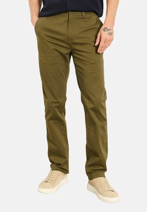 Mann trägt olivgrüne Chinos und beige Sneakers, steht mit einer Hand in der Tasche und der anderen Seite am Körper.