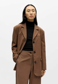 Bruine blazer met een ontspannen pasvorm, twee knopen en een zak. Gecombineerd met een zwarte coltrui en bijpassende bruine broek. Gladde stofstructuur.