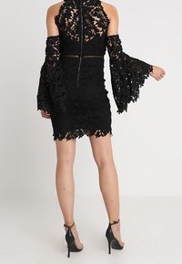 Robe en dentelle noire avec de longues manches évasées et des motifs floraux. La robe présente une silhouette ajustée et un ourlet festonné.