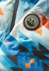 Tela estampada colorida que presenta diseños geométricos en azul, naranja y negro; incluye un parche redondo de cuero marrón con un logo de montaña.
