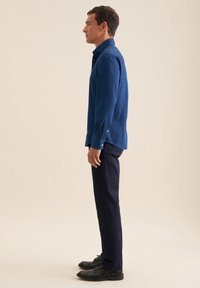 Uomo in profilo che indossa una camicia blu con bottoni, pantaloni scuri e scarpe eleganti nere, su uno sfondo beige uniforme.