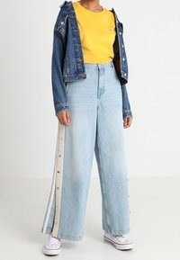 Veste en denim avec des boutons, t-shirt jaune et jeans larges bleu clair avec des détails de boutons sur le côté et une coupe décontractée. Baskets blanches.
