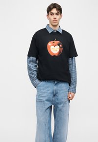 Mladý muž v čiernom tričku s veľkou oranžovou grafikou jablka cez modrú kockovanú košeľu a voľné svetlomodré rifle, stojaci s rukami v vreckách.