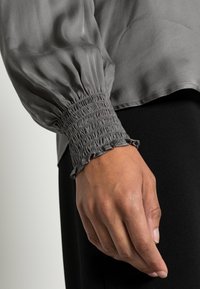 Blusa grigia in seta con maniche a sbuffo e polsini arricciati. Caratterizzata da una consistenza morbida e un bordo frangiato al polsino. Sfondo nero.