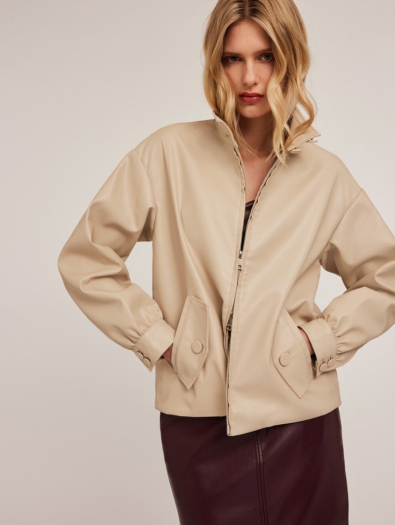Giacca in finta pelle beige con collo alto, chiusura con zip e polsini elasticizzati. Presenta tasche a toppa oblique e una texture liscia.