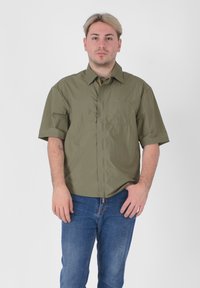 Camicia a maniche corte in verde oliva, realizzata in materiale leggero, con chiusura a bottoni sul davanti e una tasca a toppa sul lato sinistro.