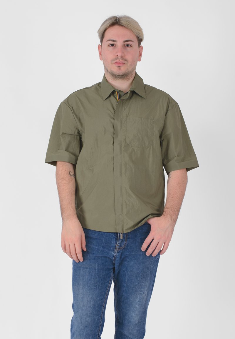 Camicia a maniche corte in verde oliva, realizzata in materiale leggero, con chiusura a bottoni sul davanti e una tasca a toppa sul lato sinistro.