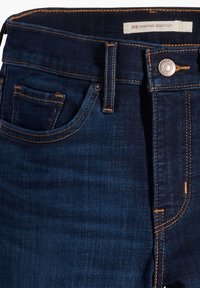Jean bootcut bleu foncé avec une texture lisse, ornés de coutures orange, d'une poche avant et d'une fermeture à bouton métallique à la taille.