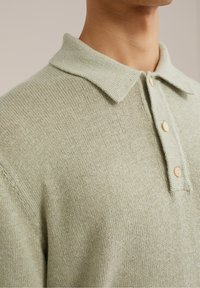 Ljust grön stickad pikétröja med krage, tre bruna knappar och ett strukturerat tyg. Närbild på krage och axelområde.