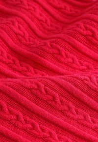 Piros kötött textil, amely dombornyomott fonott mintával, bordázott vonalakkal és puha, bolyhos szálakkal rendelkezik. Az anyag vastagnak és melegnek tűnik.