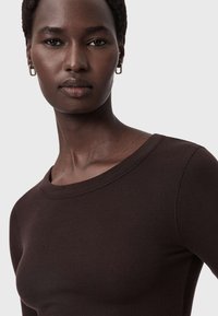 Bruine geribbelde top met lange mouwen en een ronde halslijn, gedragen door een persoon met een kort kapsel, met een gladde huid en subtiele oorbellen.
