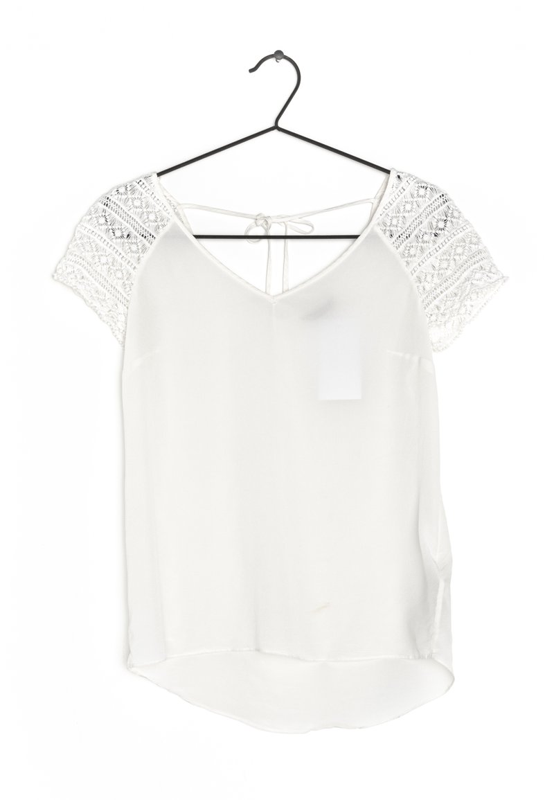 Blusa bianca con maniche a uncinetto corte, scollo a V e dettaglio di fiocco sul retro. Orlo leggermente curvo con un tessuto leggero e morbido.