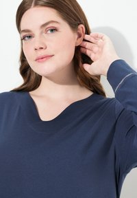 Mørkeblå, langærmet trøje med bred v-hals og blød tekstur. Funktioner en subtil udsmykning på ærmet, som komplementerer det overordnede design.