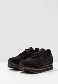 Černé tenisky s texturovaným svrškem, šněrováním a polstrovaným límcem. Mají kontrastní hnědý pruh na mezipodešvi a vzorovanou gumovou podešev.