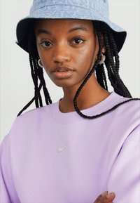 Sweatshirt roxo de gola redonda com um pequeno logótipo da Nike em prata, combinado com um chapéu de balde com padrão azul claro e cabelo trançado.