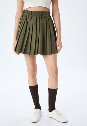 Olijfgroene plisséskirt met een rafelige zoom, een elastische tailleband en een gestructureerde stof. Gecombineerd met witte schoenen en bruine knie-sokken.