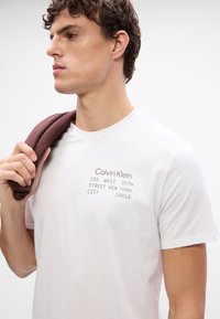 Tricou alb cu mânecă scurtă, prezentând text imprimat cu marca "Calvin Klein" și detalii de adresă; modelul ține un obiect maronii peste umăr.