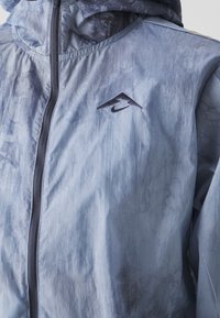 Lettvægtet hættetrøje i blå-grå tie-dye mønster. Har en frontlynlås og lille mørk logo på brystet. Glat, blank tekstur.