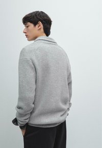 Grå ribbade stickade tröja med sjalkrage, långa �ärmar och avslappnad passform, tillsammans med mörka byxor. Diskreta texturdetaljer är tydliga.