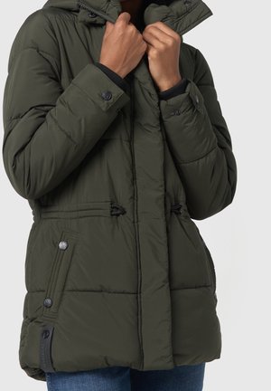 Personne ajustant le col d'une veste d'hiver matelassée vert foncé avec des boutons-pression et des poches latérales zippées, portant un jean bleu.
