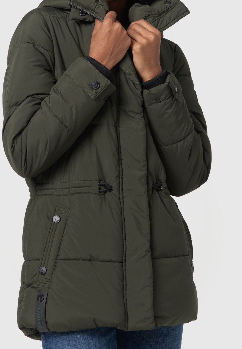 Personne ajustant le col d'une veste d'hiver matelassée vert foncé avec des boutons-pression et des poches latérales zippées, portant un jean bleu.